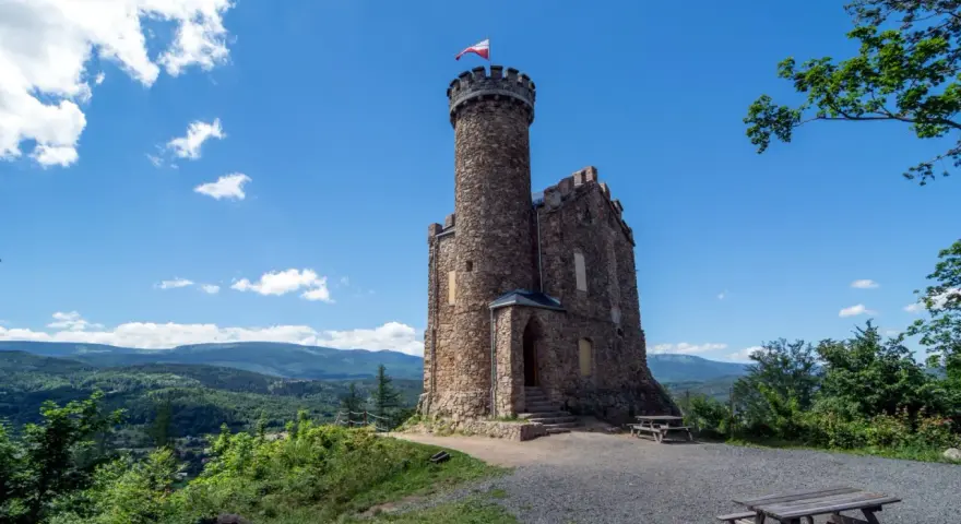 Kamienny zamek księcia Henryka z wieżą i polską flagą, otoczony zielonymi wzgórzami i błękitnym niebem.
