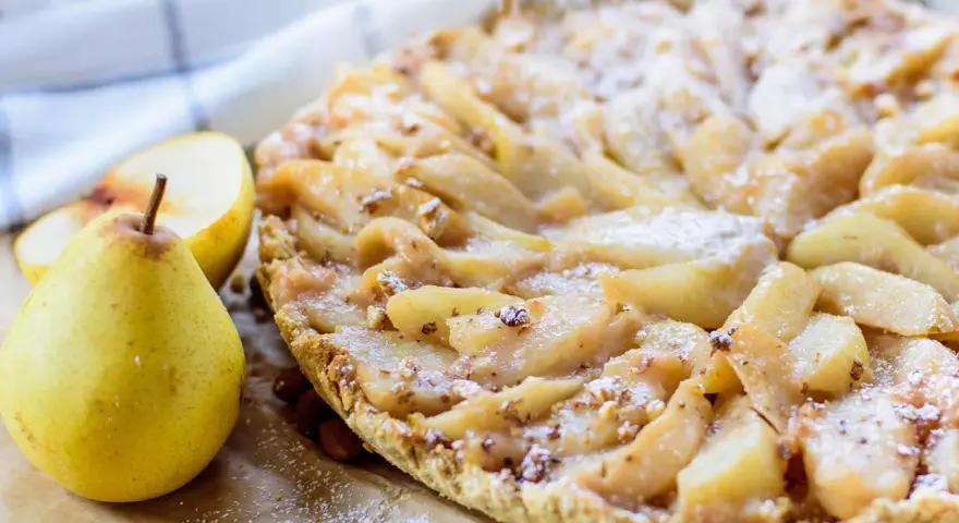 Délicieuse tarte aux poires, saupoudrée de sucre glace, avec des amandes et des poires fraîches à côté.