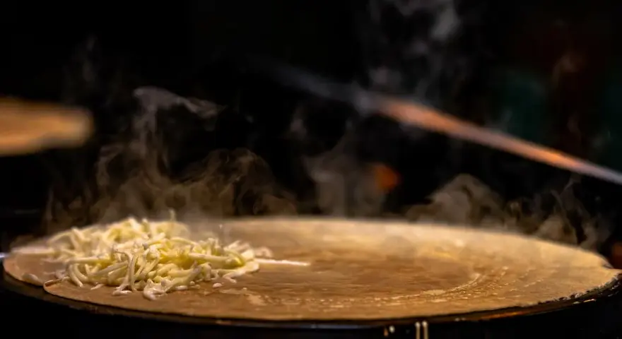 Fromage râpé fondant sur une crêpe chaude, vapeur s'élevant. Une tâche de nettoyage crêpière Tefal brûlée est visible sur le bord.