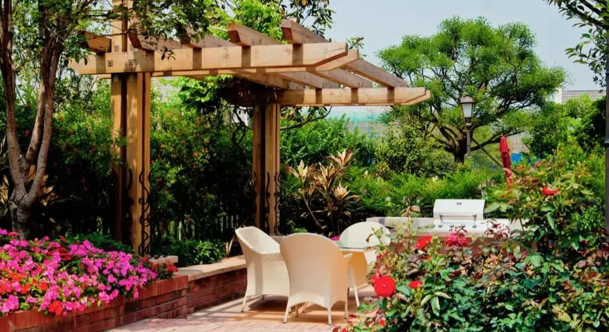 Jardín con pérgola de madera, mesa y sillas, flores rosas y rojas, y barbacoa.
