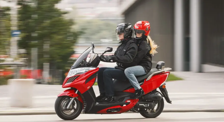 Para jadąca na czerwonym skuterze 125, ubrana w kurtki i kaski, cieszy się jazdą.