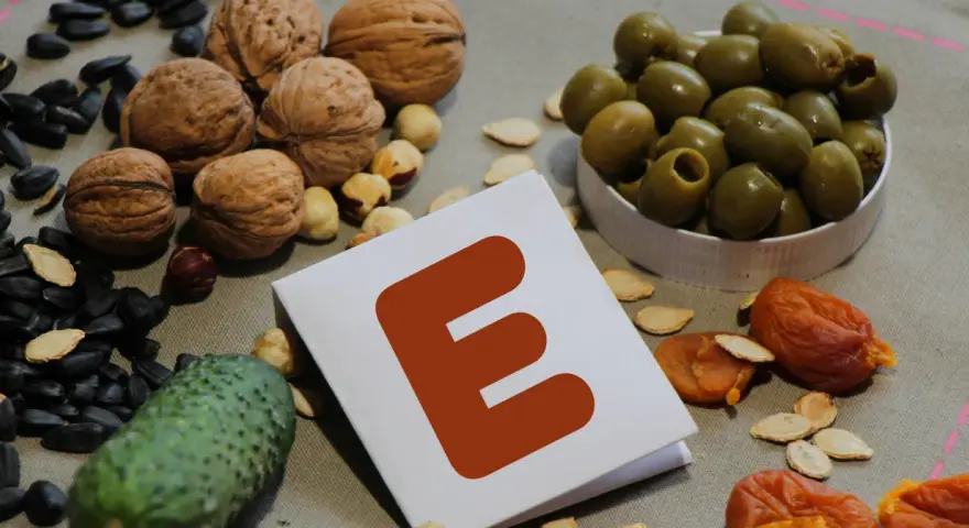 Orzechy, pestki, oliwki i suszone morele wokół kartki z literą "E" symbolizują bogactwo witaminy E.