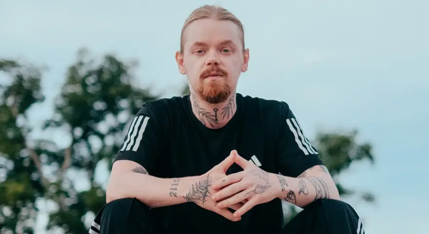 Mężczyzna z rudą brodą i blond włosami, ubrany w czarną koszulkę z trzema paskami. Na szyi i rękach widoczne tatuaże, w tym klucz i napis "kuqe 2115".