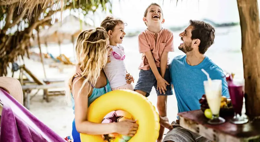 Lachende Familie am Strand mit gelbem Schwimmring und Getränken.