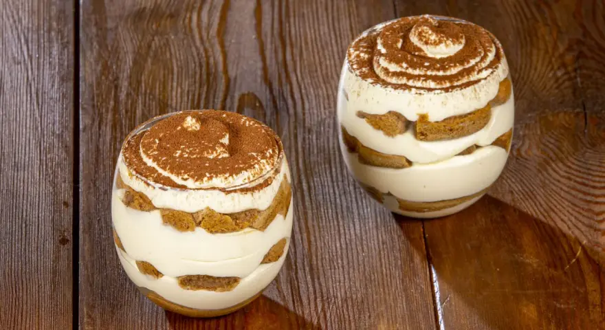 Due bicchieri di tiramisù senza uova, stratificati con crema e savoiardi, spolverati di cacao su un tavolo di legno.