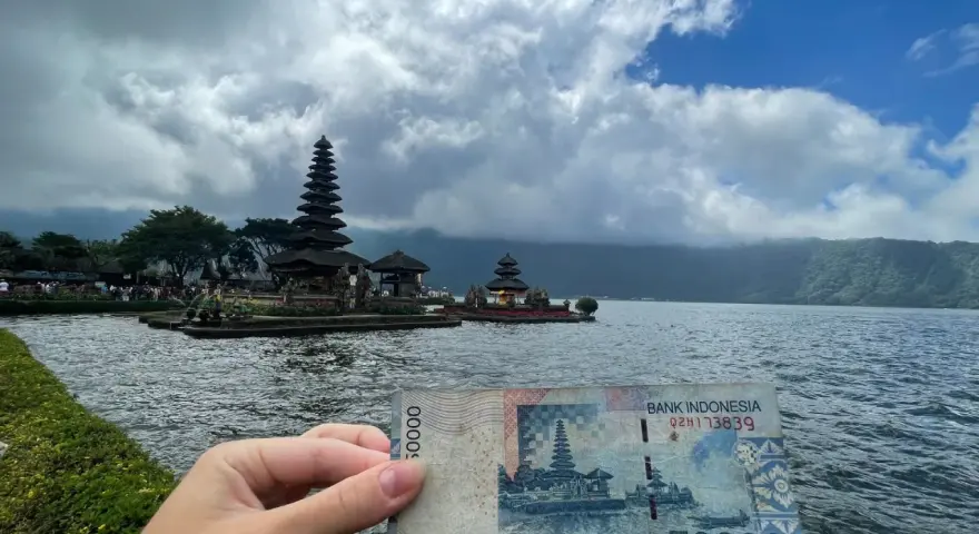 Ręka trzyma banknot 50 000 rupii na tle świątyni na wodzie. Czy Indonezja jest bezpieczna? Tak, podróżowanie po niej jest bezpieczne.