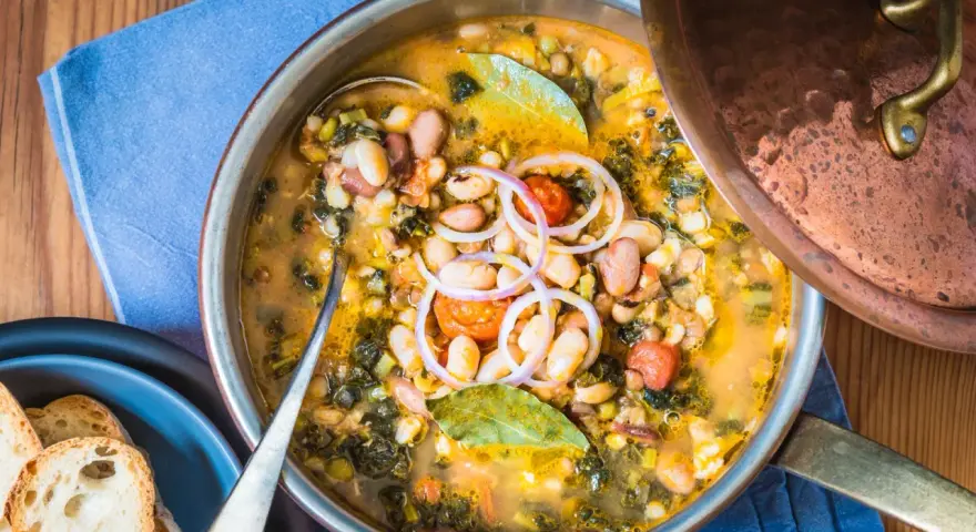 Zuppa ricca con fagioli, verdure e spezie, servita in una pentola di rame. Un piatto confortante di acqua cotta, guarnito con cipolla rossa e alloro.
