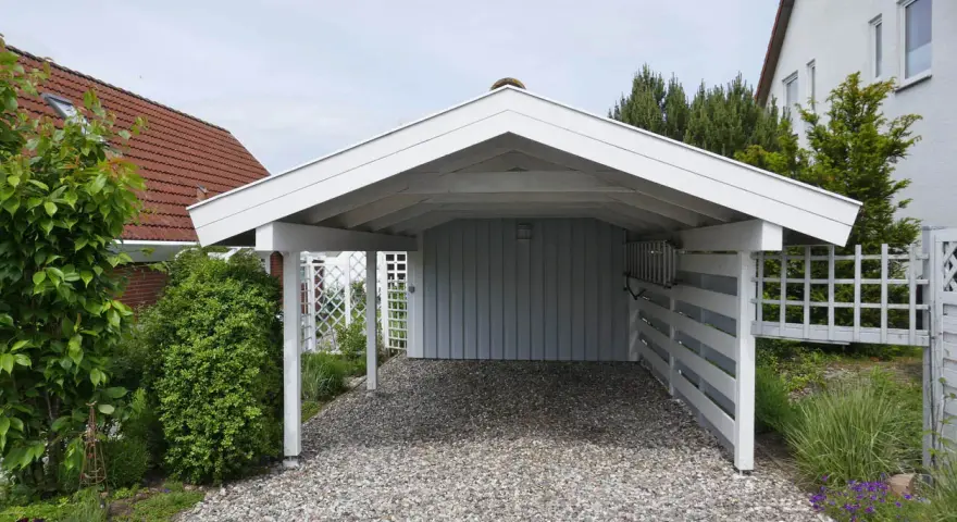 A white wooden carport with a gravel driveway, surrounded by greenery and a white lattice fence. This image shows a finished carport, a great example of how to build a carport.