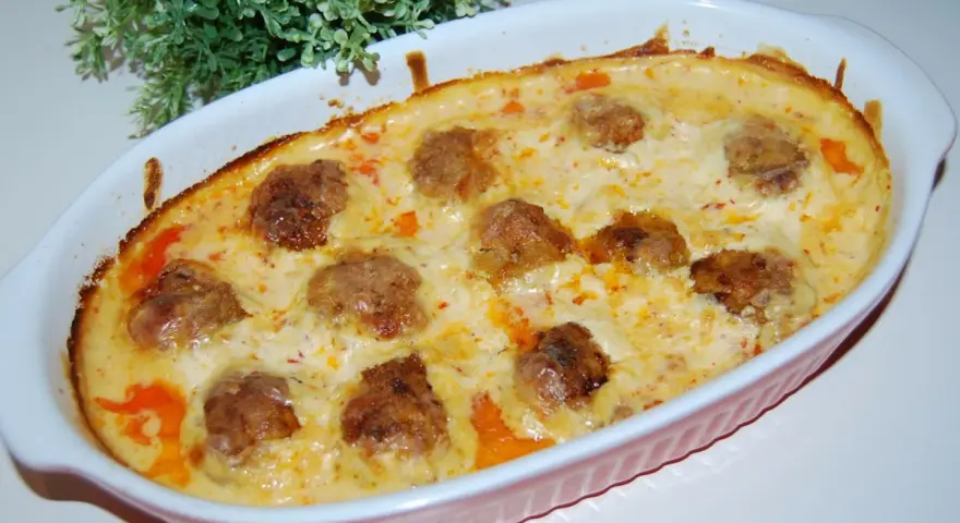 Pulpety w sosie śmietanowo-serowym, zapieczone w naczyniu, z zieloną dekoracją w tle.