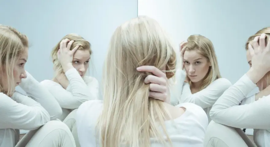 Blondynka w białej bluzie, zmagająca się z kwetaplex, widzi swoje odbicia w lustrze, każde z innym wyrazem twarzy.