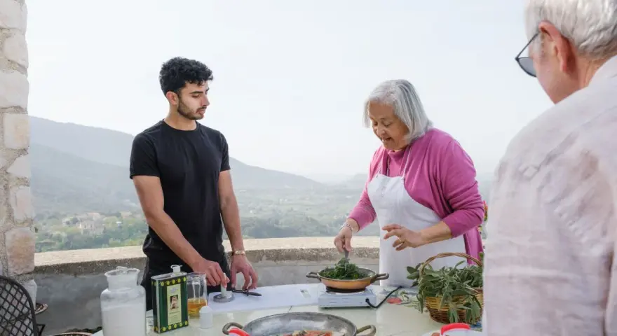 An older woman in an apron cooks greens while a young man and an older man stand nearby on a terrace with a scenic view.
