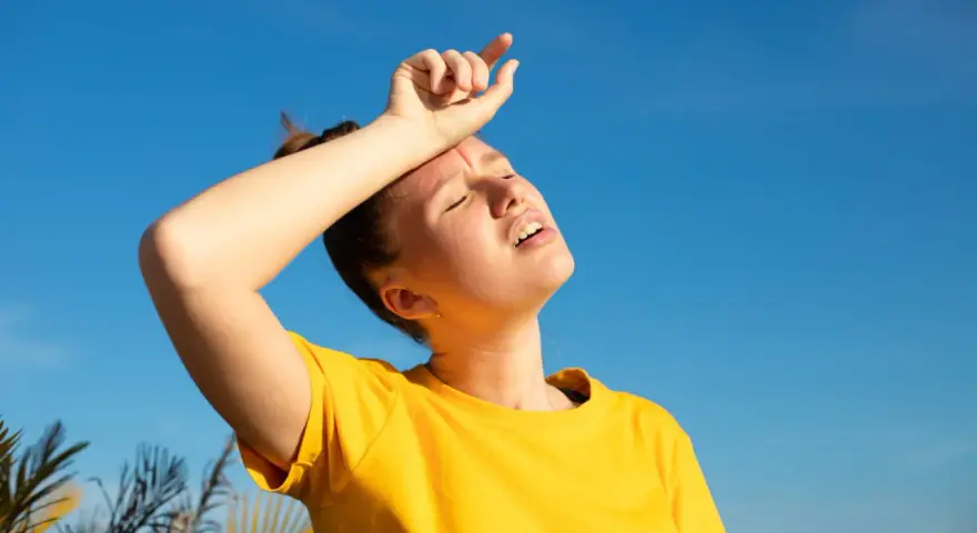 Une jeune femme en t-shirt jaune, le visage marqué par la chaleur, essuie son front sous un ciel bleu intense. La durée d'une insolation est palpable.