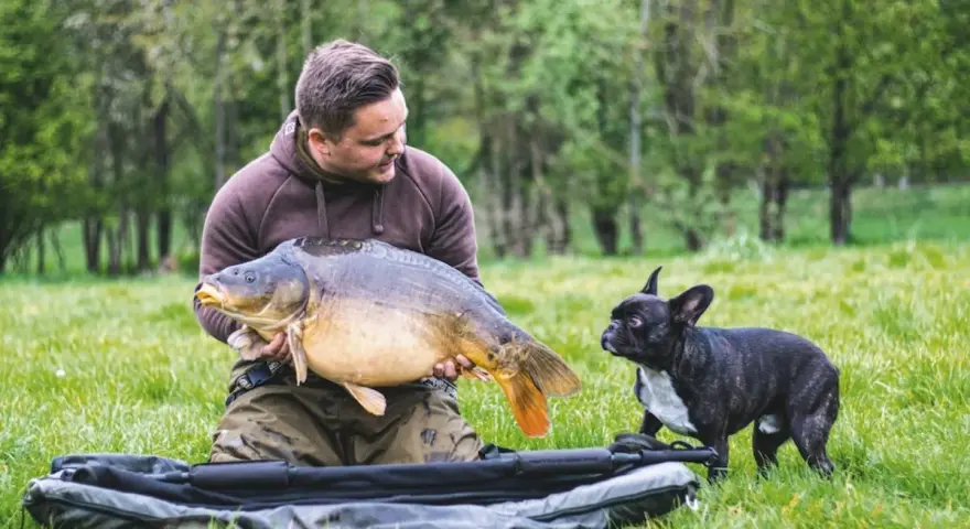 Ein Angler präsentiert stolz einen großen Karpfen, während sein kleiner Hund neugierig zusieht. Ein erfolgreicher Tag beim feedern auf Karpfen.