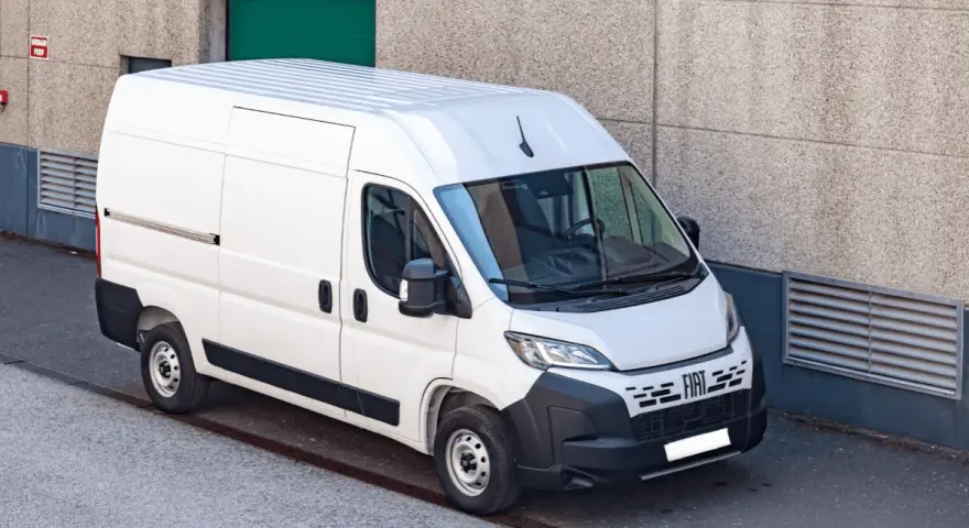 Biały bus dostawczy Fiat Ducato zaparkowany przy betonowej ścianie budynku.