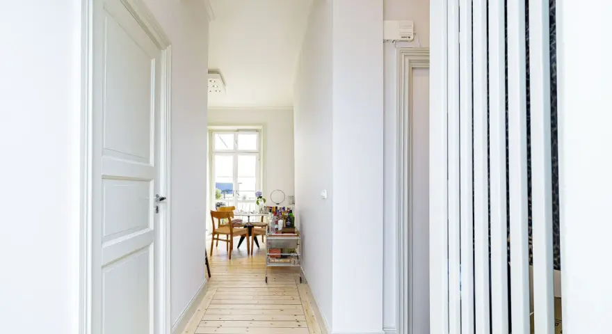 Ljus hall med trägolv och vita dörrar, perfekt för en renovera hall. Genom en valvöppning syns ett runt bord med stolar och ett fönster.
