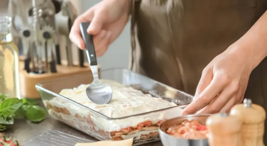 Osoba nakłada sos na warstwy makaronu i mięsa, przygotowując do pieca lasagne.