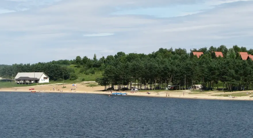 Plaża nad jeziorem Chańcza tętni życiem. Ludzie odpoczywają na piasku, kąpią się w wodzie, a w tle widać domki i las.