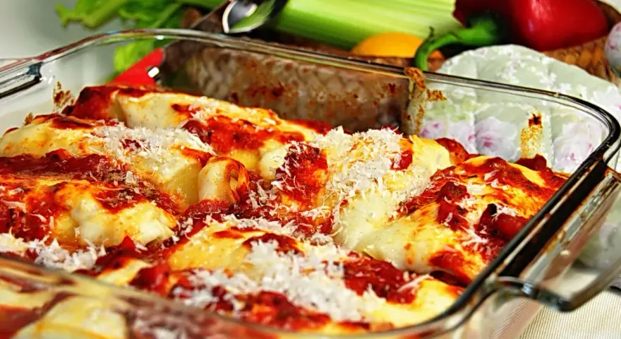 Pyszne, zapiekane cannelloni w sosie pomidorowym, posypane parmezanem. Obok świeże warzywa, idealne do dania.