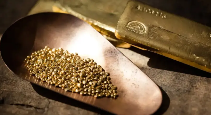 Piccoli granelli scintillanti di oro riciclato riposano in una paletta di rame, accanto a lingotti dorati.