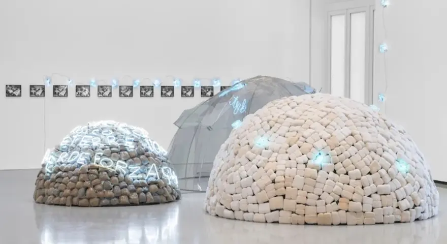 Installation d'art povera : dômes en pierres et en tissu, ornés de néons bleus, sous une structure transparente.