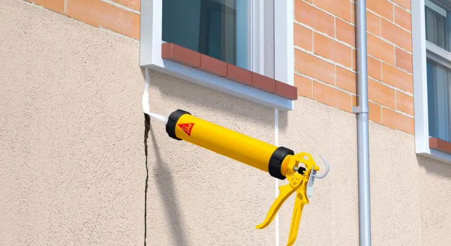 Pistola aplica sellador blanco en una grieta del rebozado de la pared exterior, junto a una ventana.