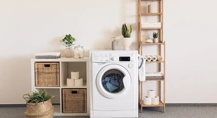 Lavadora blanca con cesta de mimbre, plantas y accesorios que se colocan en una prenda.