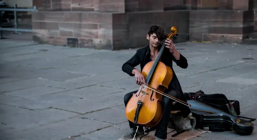 Ein Mann spielt auf einem Cello-Musikinstrument vor einer Steinfassade.