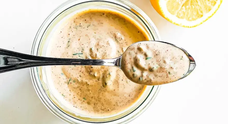 En sked krämig remouladsås med örter lyfts ur en burk. Bredvid ligger en halv citron. Perfekt för ditt remouladsås recept!