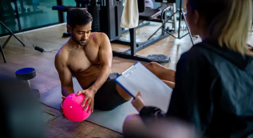 Man med rosa boll på gymmatta, tränare med clipboard. De följer sitt gymprogram.