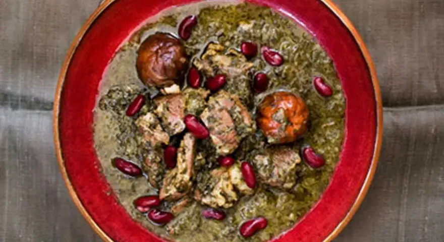 En skål med ghorme sabzi, en mustig persisk gryta med kött, örter och röda bönor, serverad i en röd kantad skål.