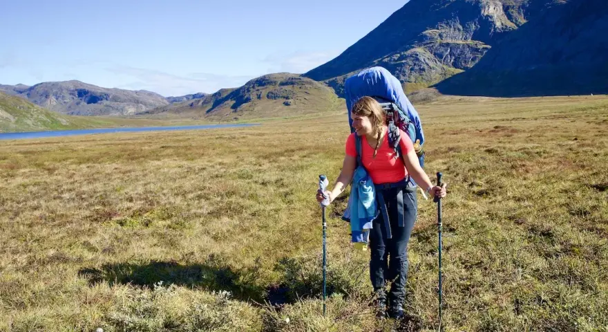 Frau wandert mit großem Rucksack und Stöcken über einen grasbewachsenen Hügel. Im Hintergrund ein See und Berge. Ein Abenteuer auf dem Arctic Circle Trail.