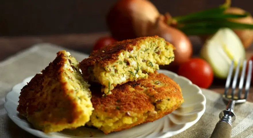 Pyszny kotlet jajeczny z ziołami, podany na talerzyku. W tle cebula i pomidorki.