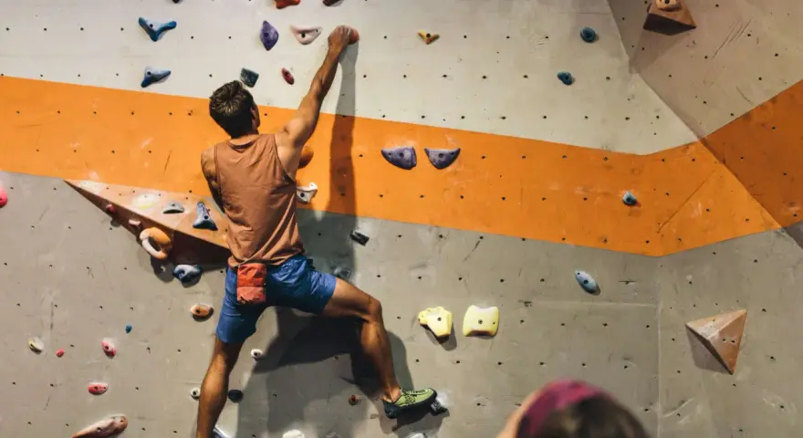 Mężczyzna wspina się po ścianie z kolorowymi chwytami. To właśnie bouldering – wspinaczka bez liny na niskich ściankach.