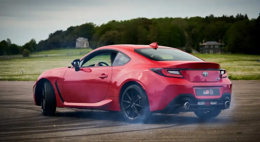 Czerwona Toyota 86 GR w poślizgu, z opon dymiących na asfalcie. W tle zielone pola i drzewa.