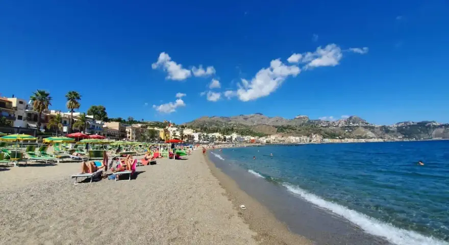 Spiaggia affollata a Giardini di Naxos, con lettini, ombrelloni e persone che si rilassano sotto il sole siciliano.