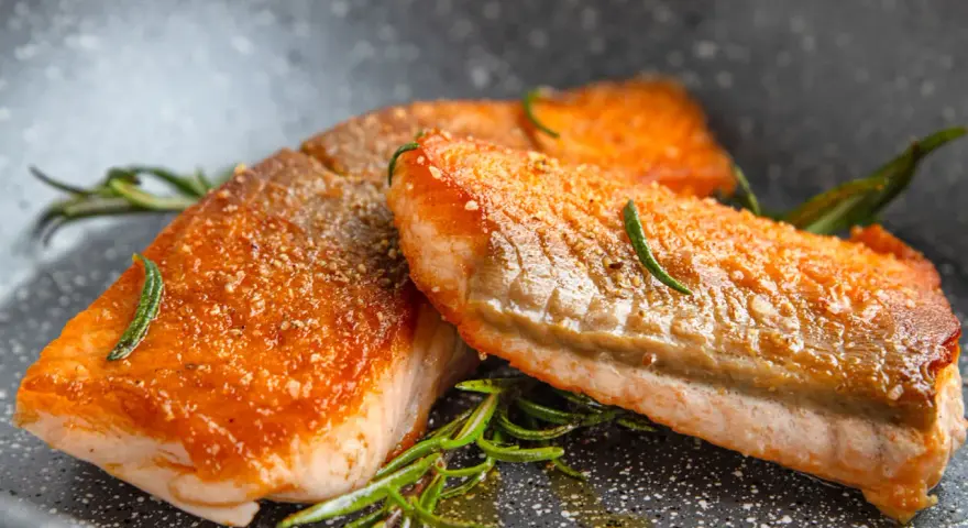Zwei saftige Lachsfilets mit knuspriger Haut, perfekt gebraten, liegen in einer Pfanne mit Rosmarin und Öl.
