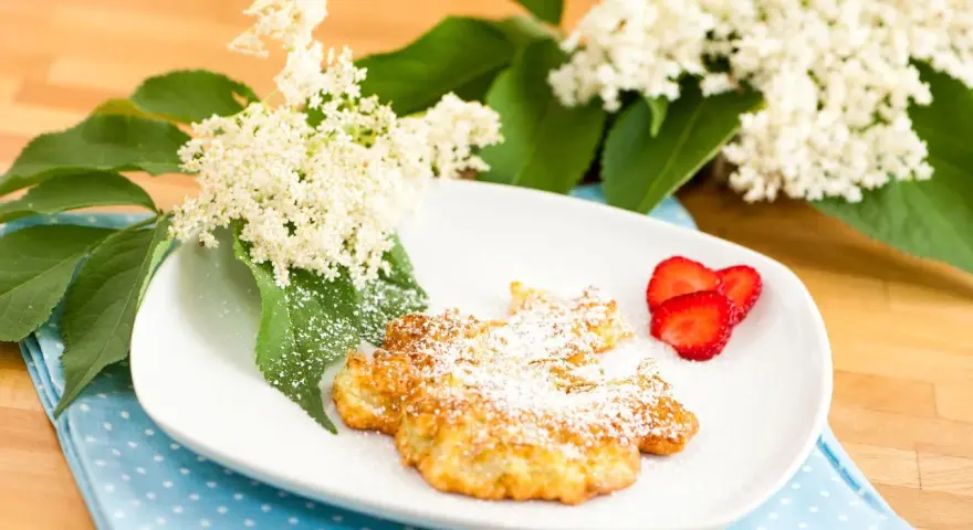 Fiori di sambuco fritti, spolverati di zucchero a velo, serviti su un piatto bianco con fragole fresche e decorati con fiori di sambuco.