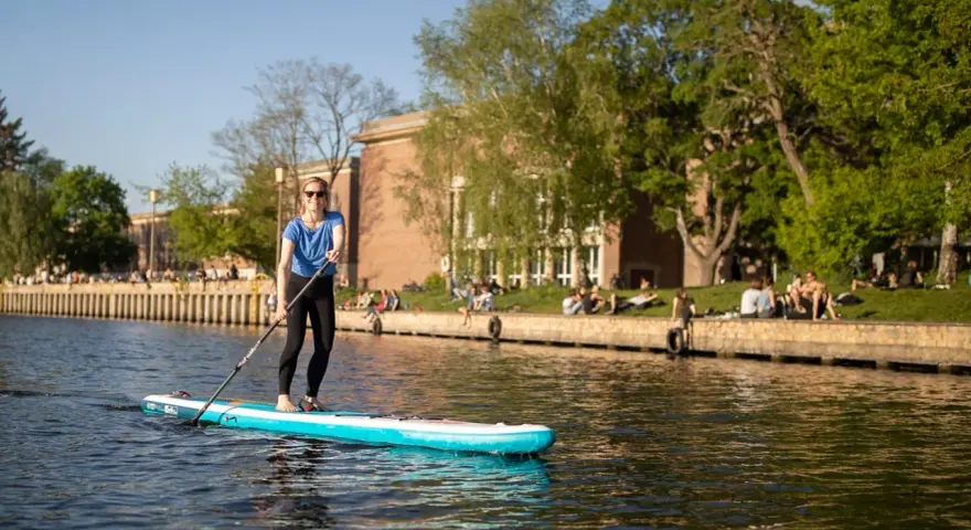 Frau genießt Stand Up Paddling auf einem Fluss, während Menschen am Ufer entspannen.