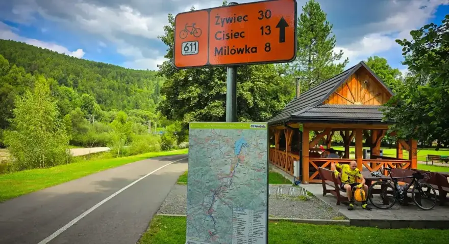 Znak szlaku rowerowego 611 Velo Soła wskazuje kierunki. Obok mapa i wiata z rowerzystą.
