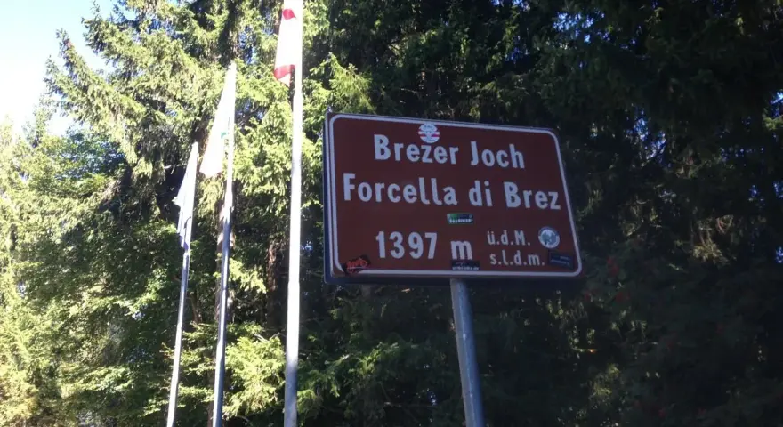 Cartello indicante "Forcella di Brez" a 1397 m, con bandiere e alberi sullo sfondo.