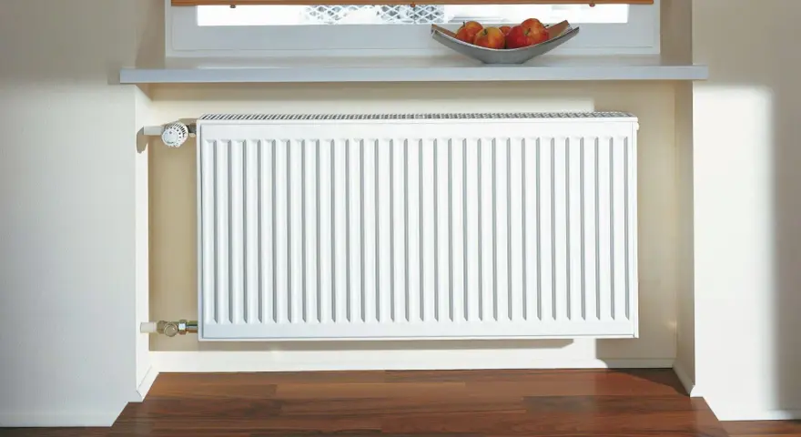 Radiador blanco de aerotermia con aletas de aluminio, bajo una ventana con persiana y un cuenco de manzanas.