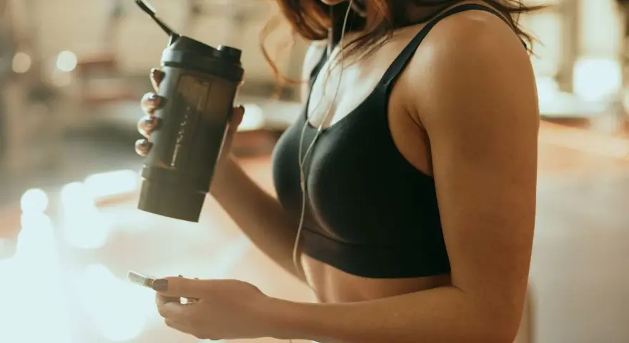 Kobieta w sportowym topie trzyma shaker, zastanawiając się, kiedy pić białko przy odchudzaniu. Słucha muzyki na telefonie.
