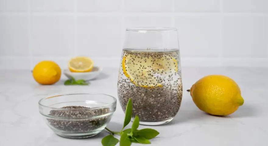 Vaso con agua de chia, rodaja de limón y semillas de chía en un bol. Limones y hojas verdes completan la escena.