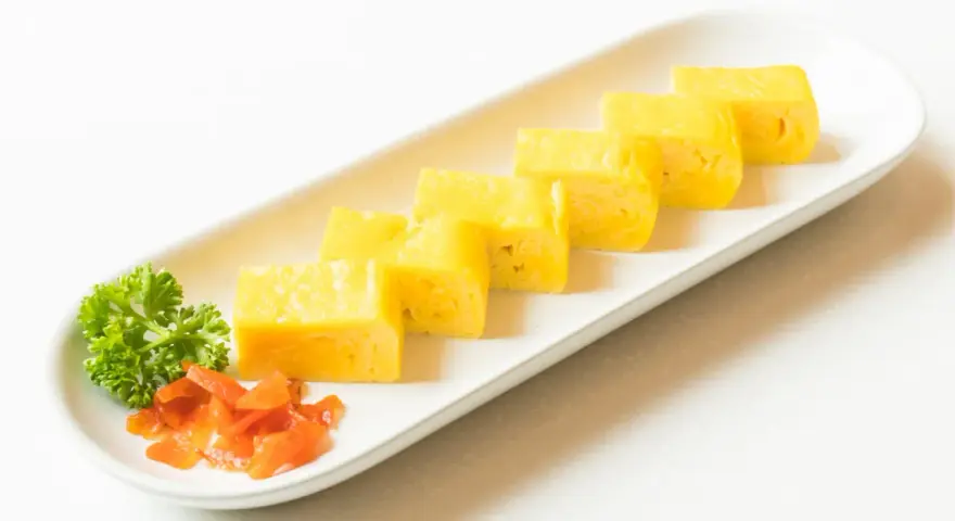 Sześć kawałków puszystego omletu tamago ułożonych na białym talerzu, z gałązką pietruszki i pomarańczowymi dodatkami.