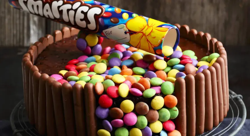 Eine einfache Geburtstagstorte für Jungs: Schokoladenkuchen, umhüllt von Schokoriegeln und bedeckt mit bunten Smarties, die aus einer aufgeschnittenen Rolle rieseln.