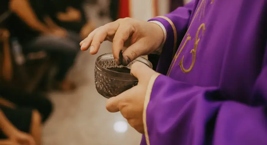 A priest in a purple robe takes ashes from a small bowl, preparing for the first day of Lent prayer.