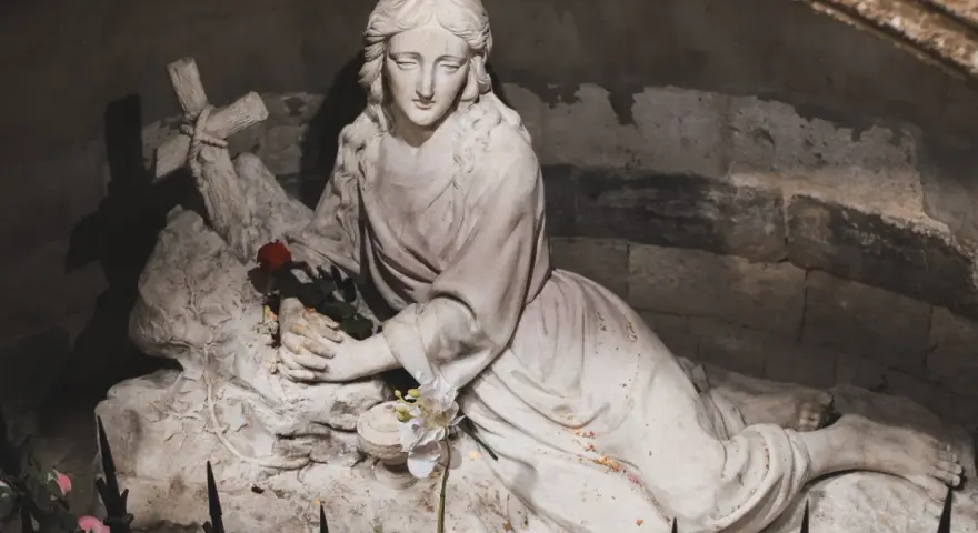 A stone sculpture depicts the serene body of Mary Magdalene, resting with a cross and a single red rose.