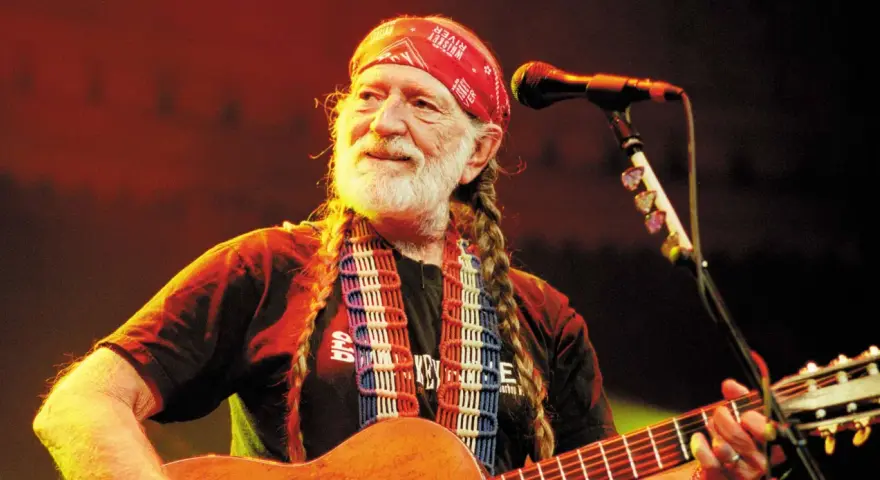 Willie Nelson mit roter Bandana und Zöpfen spielt Gitarre. Ein Mikrofon steht bereit für den Titel von Willie Nelson.
