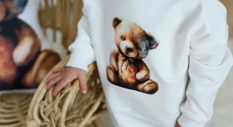 Biała bluza Moschino z uroczym misiem w stylu vintage. Dziecko trzyma się wiklinowego koszyka.