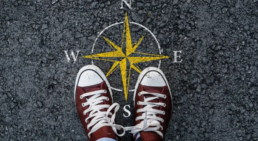 Feet stand on a compass, symbolizing the start of a journey. This image can represent the **mission creep meaning**, where initial goals expand unexpectedly.